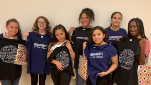 Seven teenage girls holding up and or wearing black and navy blue t-shirts with human rights slogans printed on them.